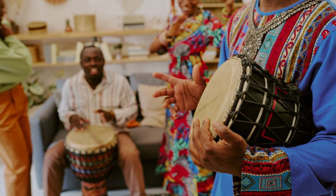 Group of Black adults celebrating Kwanzaa, playing traditional drums and dancing in living room, focus on hands of Black man drumming in foreground, joyful cultural gathering